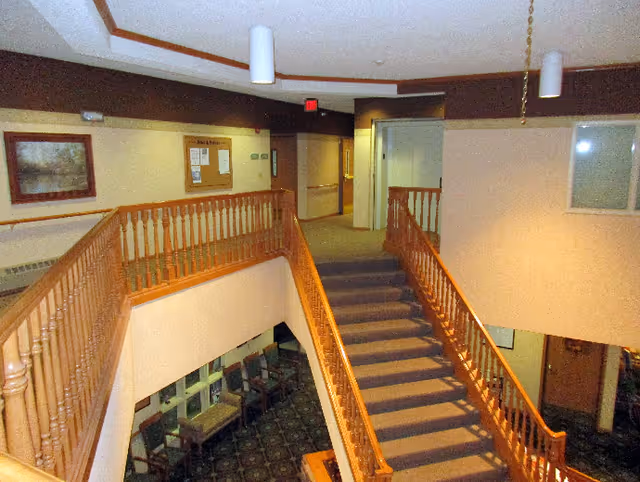 Interior view of a senior living facility showing a carpeted staircase with wooden railings leading down to a seating area with chairs and tables. The upper level has a hallway with doors and a bulletin board on the wall. The ceiling has recessed lighting fixtures.