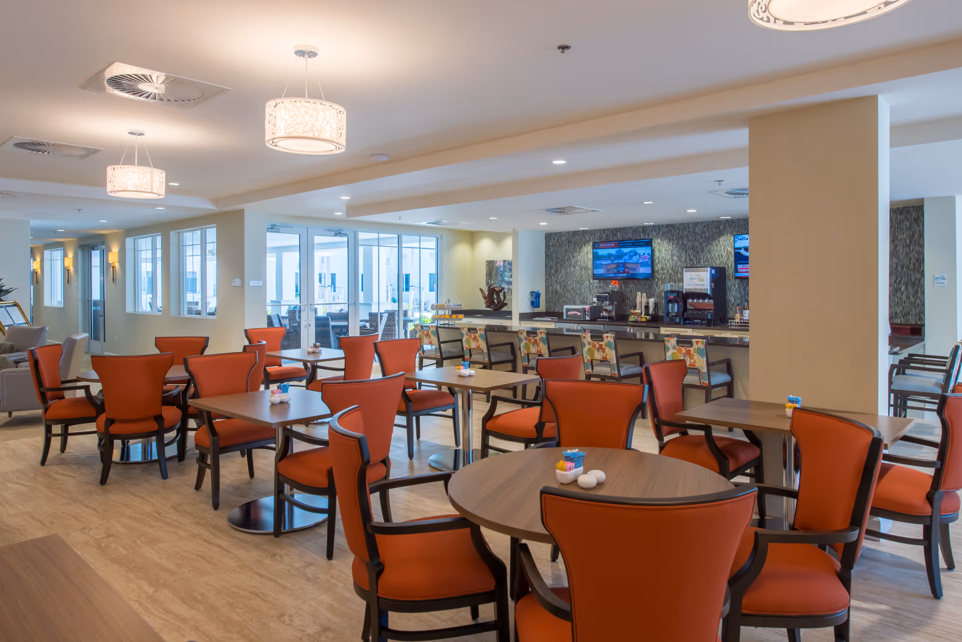 A spacious dining area with multiple wooden tables and orange cushioned chairs. The room features modern pendant lights hanging from the ceiling, a bar counter with colorful bar stools, and two televisions mounted on a textured wall. Large windows and glass doors provide natural light and a view of an outdoor seating area.