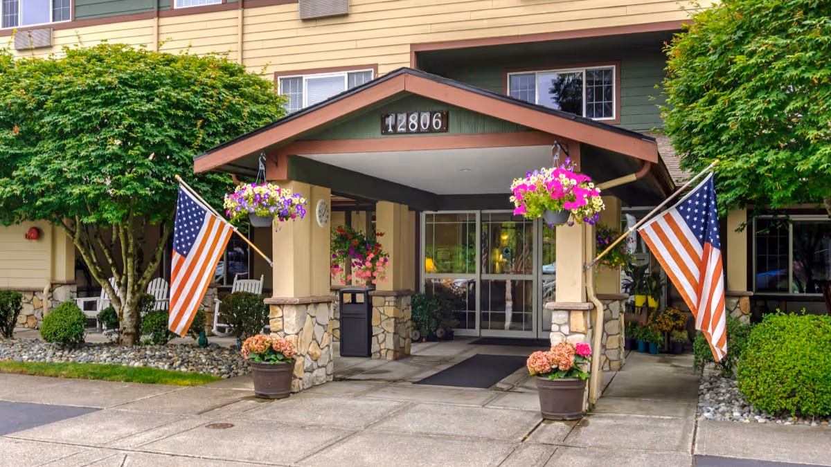 Covered entrance of a senior living building with a gabled portico showing address 12806, American flags, hanging flower baskets, and potted plants.