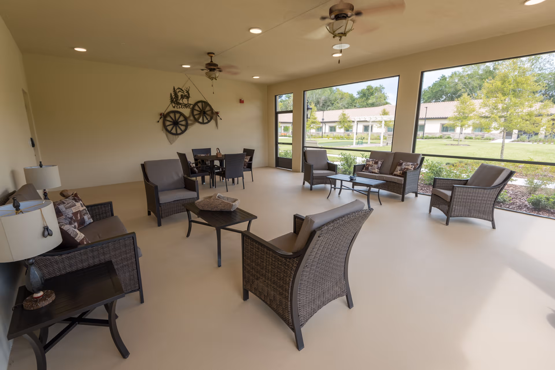 Screened outdoor seating area with wicker chairs, tables, and ceiling fans overlooking landscaped grounds.