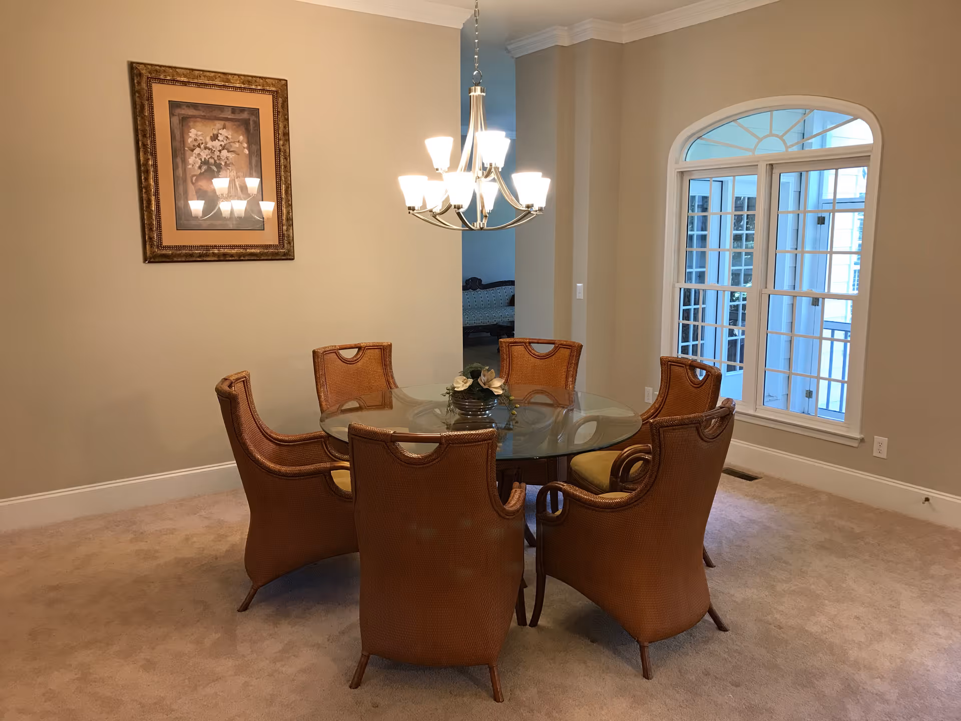 A dining room with a round glass table surrounded by six wicker chairs with cushions. A chandelier with multiple lights hangs above the table. There is a framed floral painting on the beige wall and a large window with white trim letting in natural light.