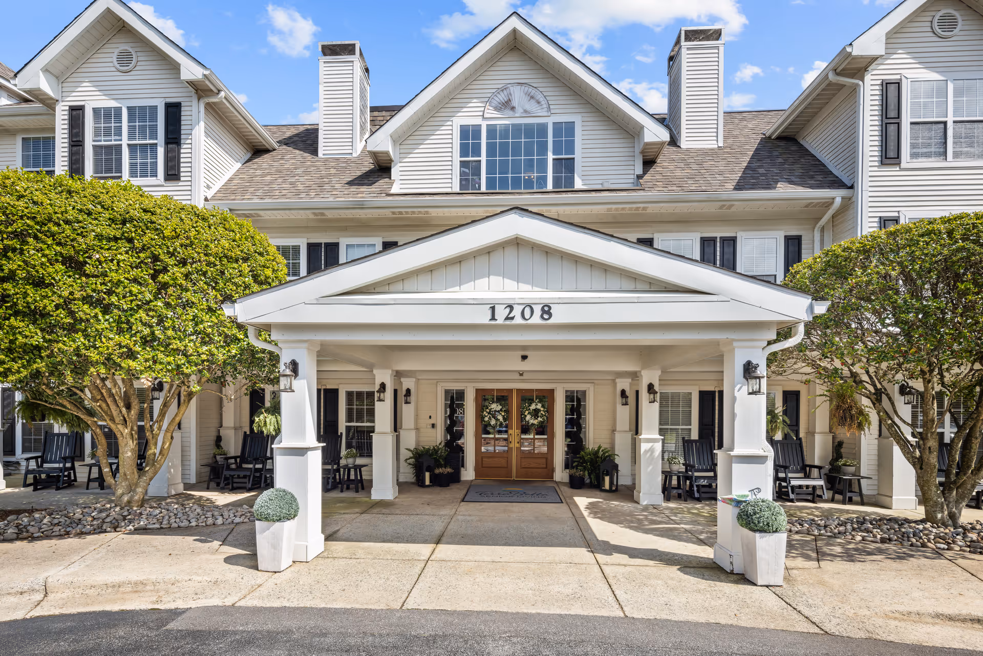Front entrance of a multi-story senior living building with a covered portico supported by columns and the number 1208 above the doors.