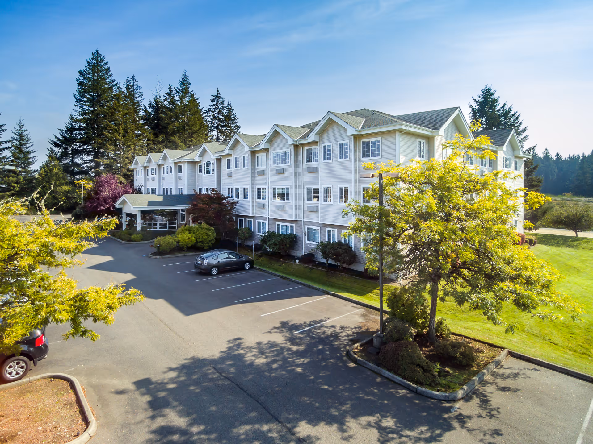 Three-story assisted living building with a covered entrance, parking lot and landscaped trees under a blue sky.