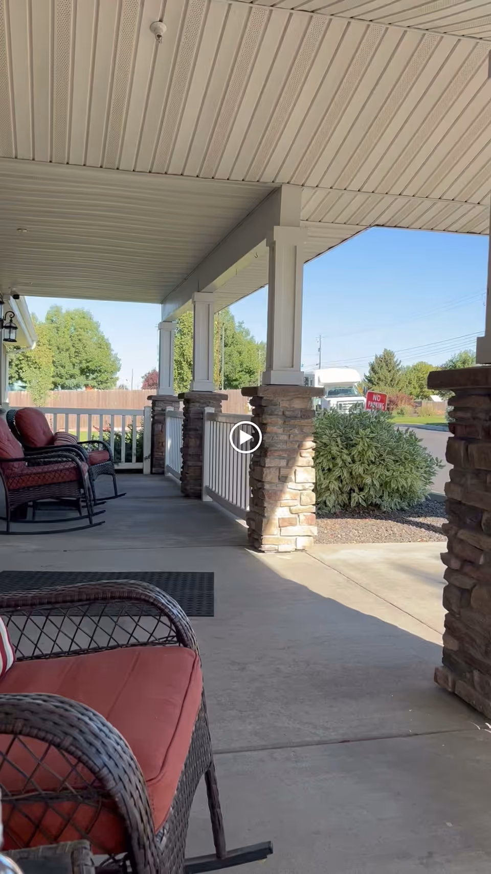 Covered outdoor porch area with wicker rocking chairs featuring red cushions, stone pillars supporting the roof, and a view of greenery and a street with a 'No Parking' sign in the background.