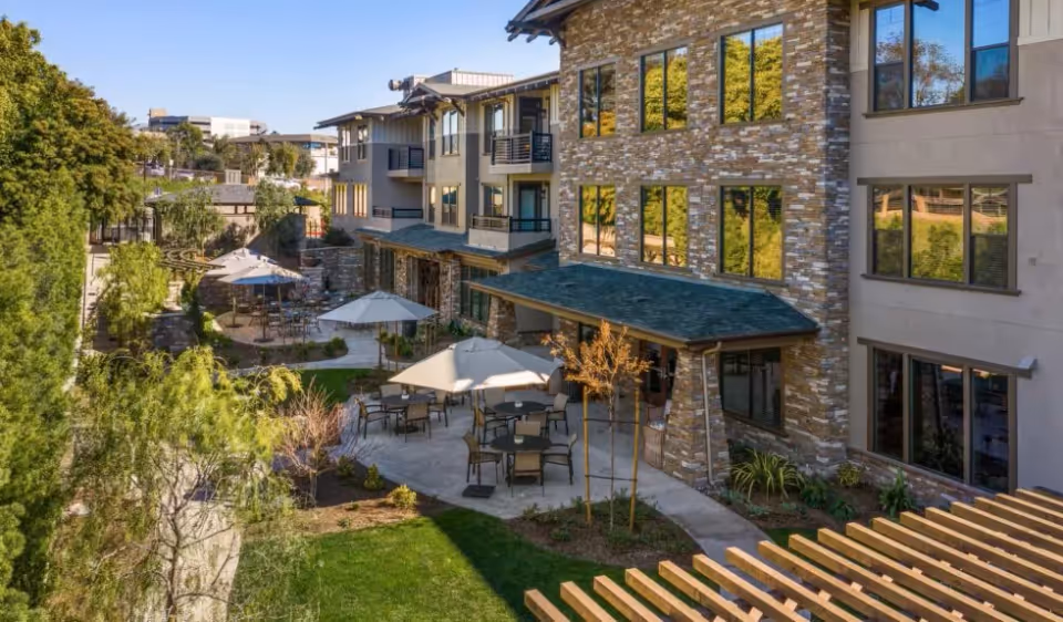 Outdoor courtyard at a senior living facility with patio tables and umbrellas beside a multi-story stone-clad building.