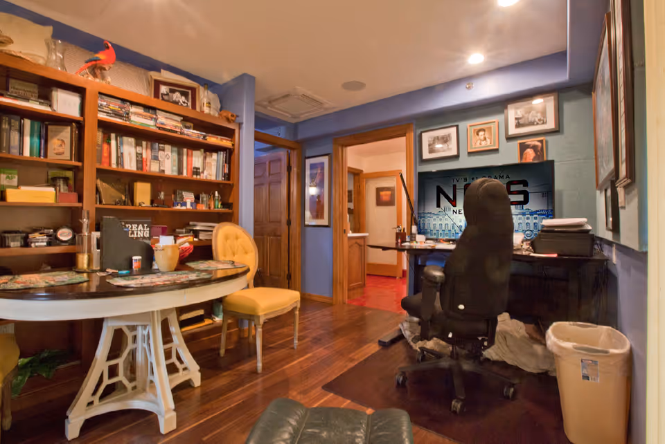 A cozy interior room with wooden flooring featuring a round table with placemats and a yellow cushioned chair. There is a large wooden bookshelf filled with books and decorative items. A black office chair faces a desk with a computer monitor displaying a news program. The walls are painted blue and adorned with framed pictures. A trash bin is placed near the desk.