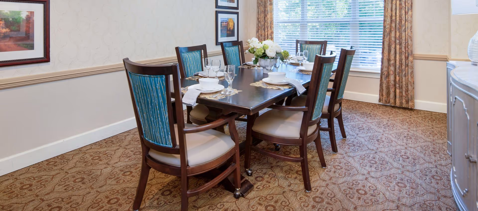 A dining room with a dark wooden table set for six people. The chairs have wooden frames with blue patterned upholstery on the backrests and beige cushions. The table is set with white bowls, plates, glasses, and silverware on woven placemats. A floral centerpiece is placed in the middle of the table. The room has beige patterned carpet, light-colored walls with framed artwork, and a window with blinds and floral curtains.