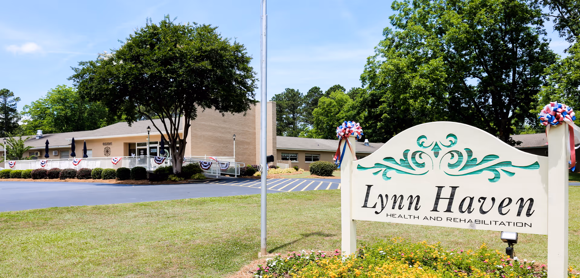 Exterior view of Lynn Haven Health and Rehabilitation facility with a large sign in the foreground surrounded by flowers and greenery. The building is a single-story structure with a beige facade, a ramp, and patriotic decorations along the railing. Trees and a clear blue sky are visible in the background.