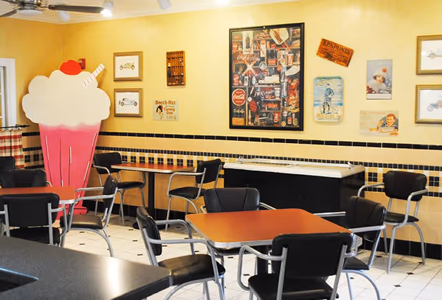 A retro-style dining area with several tables and black chairs. The walls are decorated with vintage posters and signs, including a large pink and white ice cream sundae cutout. The room has yellow walls with a black and yellow tiled border and a white tiled floor.