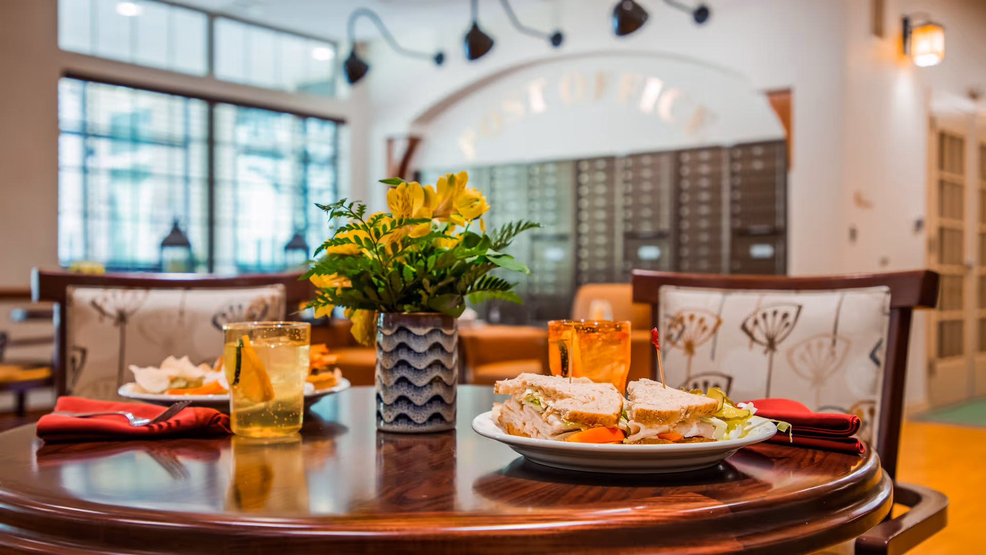 A wooden dining table set with plates of sandwiches and glasses of iced tea, with a vase of yellow flowers in the center. The background shows a bright room with cushioned chairs and a sign that reads 'Post Office'.