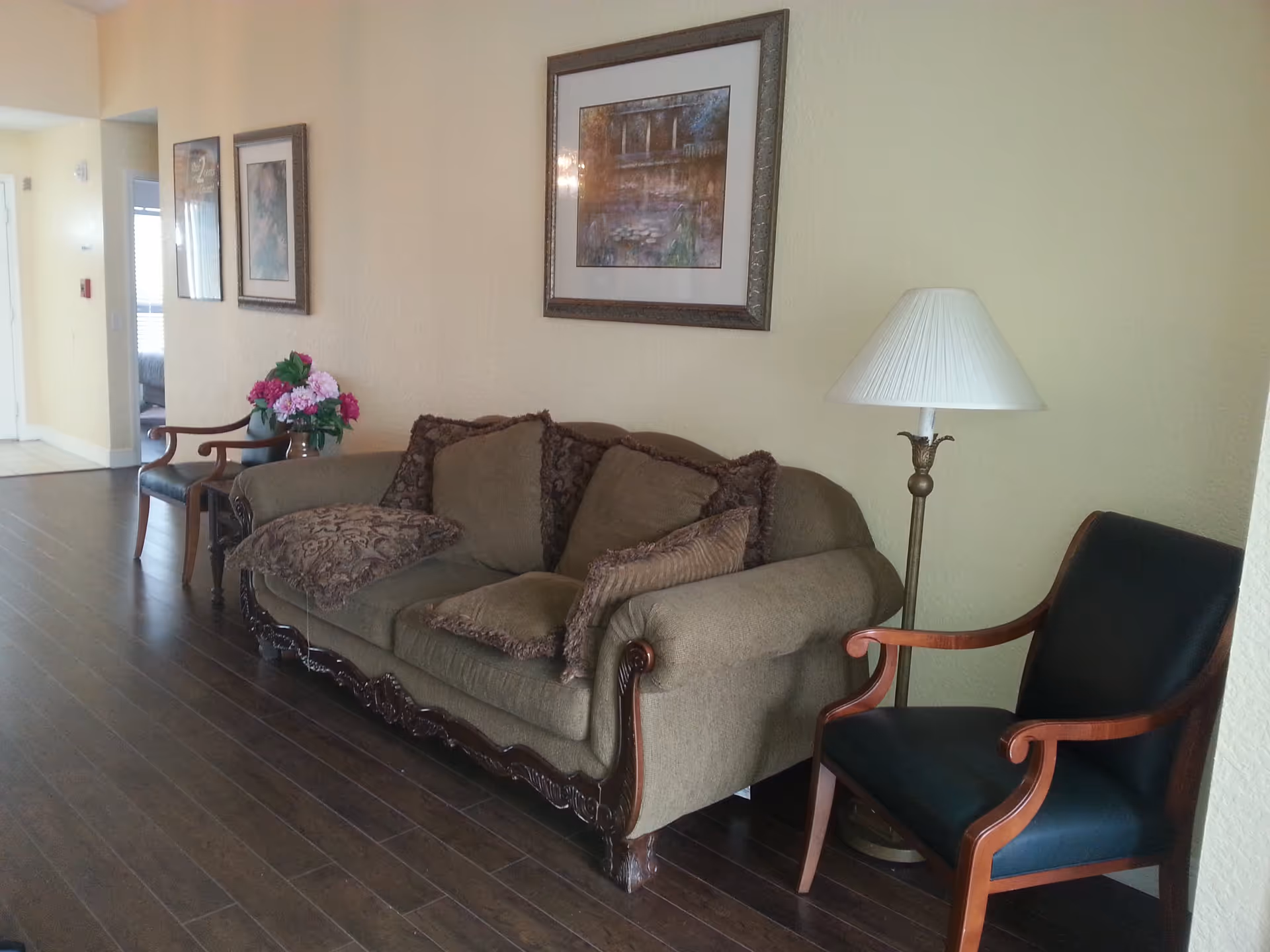 A cozy interior hallway area with a brown upholstered sofa adorned with multiple cushions, a wooden armchair with dark upholstery, a floor lamp with a white lampshade, and framed artwork on the beige wall. A small table with a vase of pink and purple flowers is placed next to another wooden chair further down the hallway.
