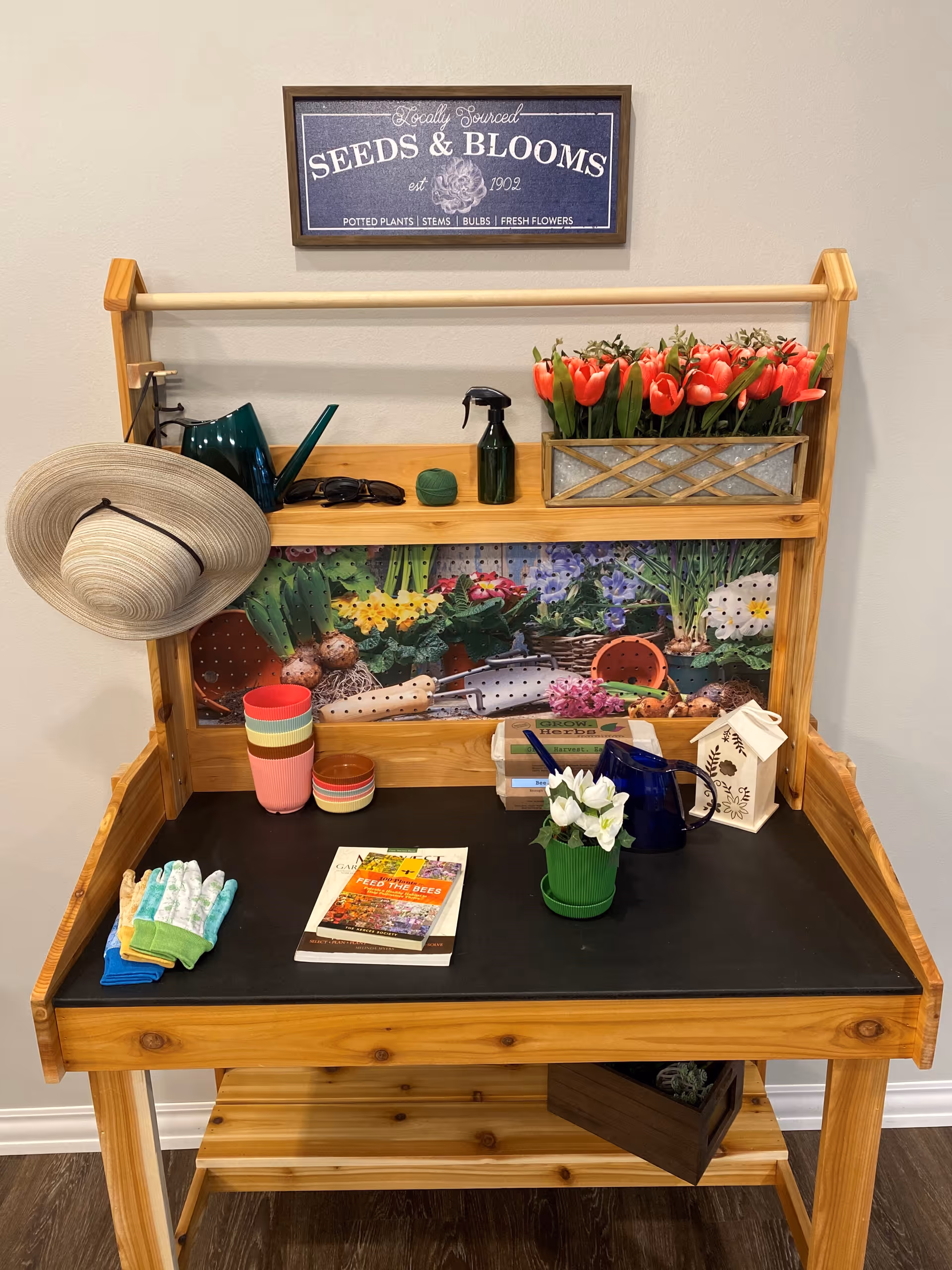 A wooden gardening potting bench with various gardening items including a straw hat hanging on the side, a green watering can, a spray bottle, a container with red tulips, gardening gloves, colorful plant pots, gardening books, a small green potted plant, a blue pitcher, and a decorative birdhouse. Above the bench is a sign that reads 'Locally Sourced SEEDS & BLOOMS est 1902 POTTED PLANTS | STEMS | BULBS | FRESH FLOWERS'.