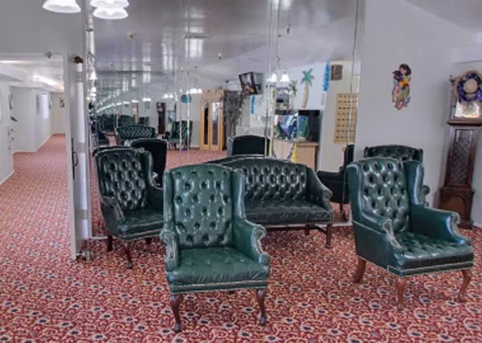 A sitting area with green leather tufted armchairs and a matching loveseat on a red patterned carpet. The room has mirrored walls reflecting the furniture and ceiling lights. There are decorative wall hangings and a grandfather clock on the right side.