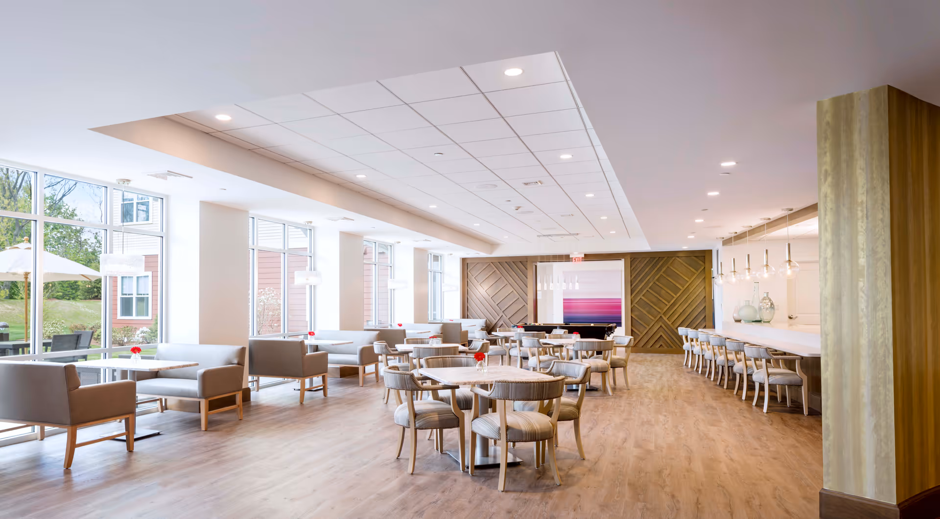 A spacious, modern dining area with multiple round tables and chairs, cushioned bench seating along the windows, and a long counter with bar stools. Large windows on the left side let in natural light and provide a view of outdoor greenery and umbrellas. The room features light wood flooring, white walls, and decorative wooden panels on the far wall.