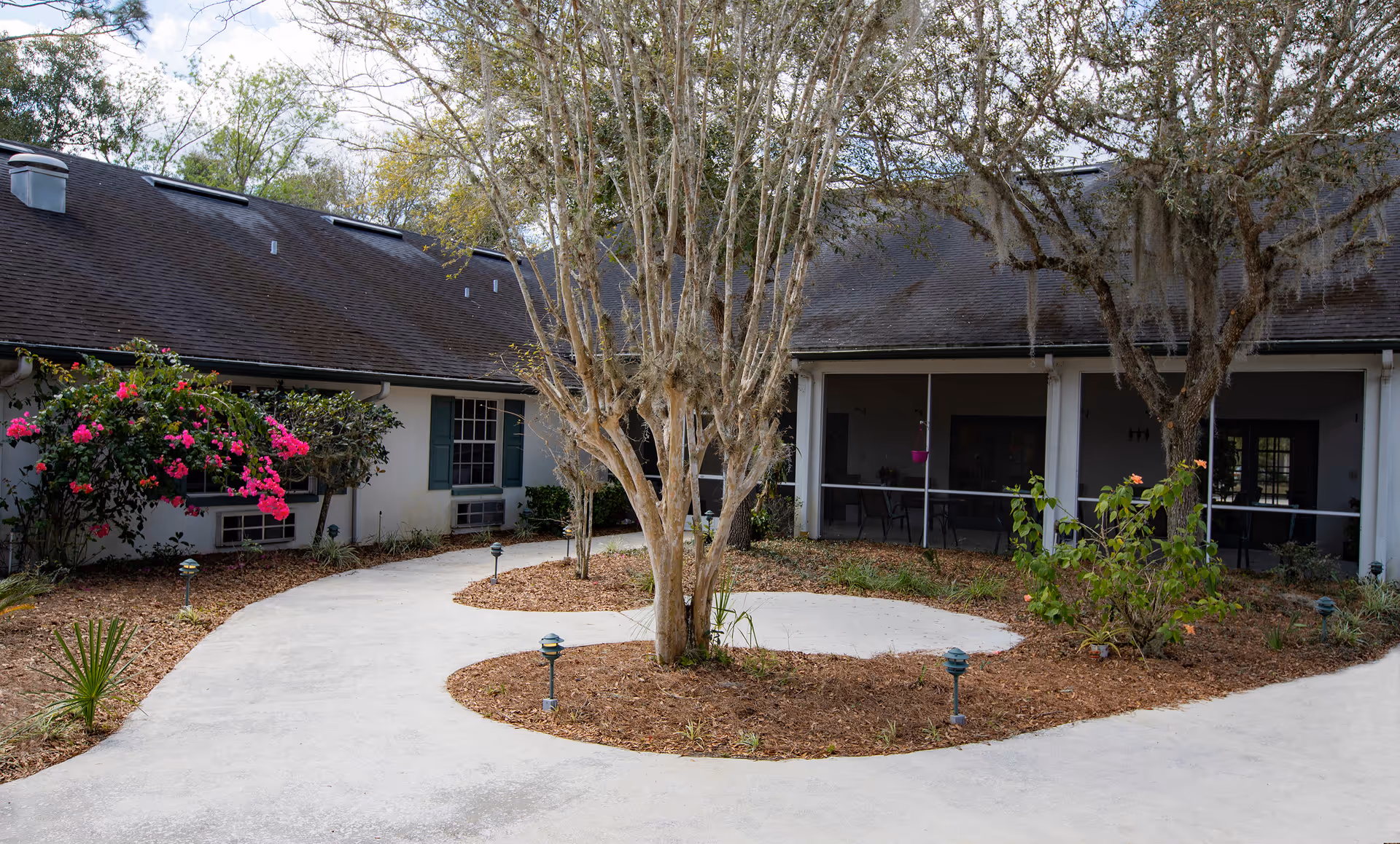 Outdoor courtyard area of a senior living facility with a curved concrete walkway, landscaped garden beds with trees and flowering bushes, and a building with a dark roof and screened porch in the background.