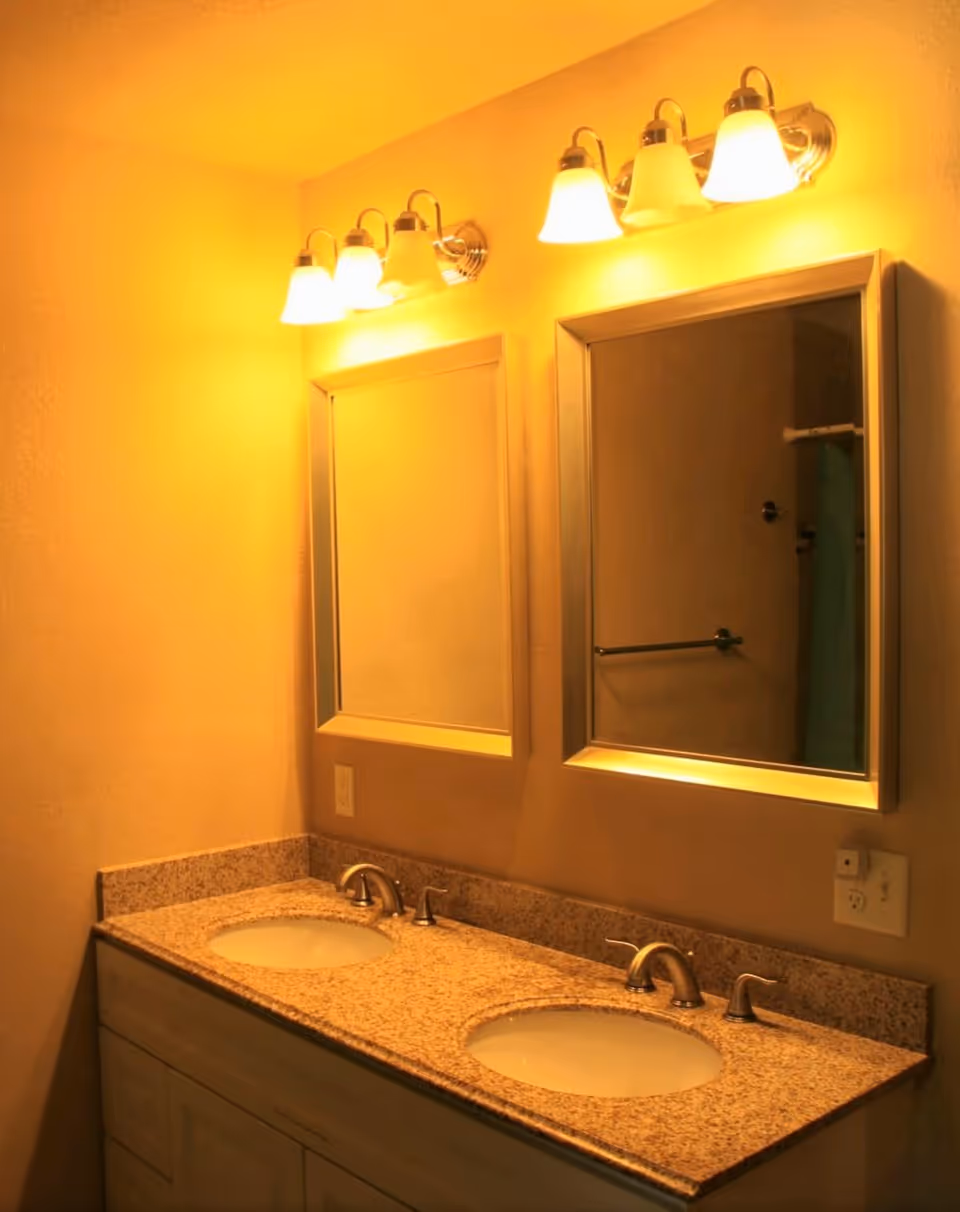 A bathroom vanity with a granite countertop featuring two sinks and two mirrors above each sink. Each mirror has a light fixture with three bulbs mounted above it. The wall is painted a warm beige color.