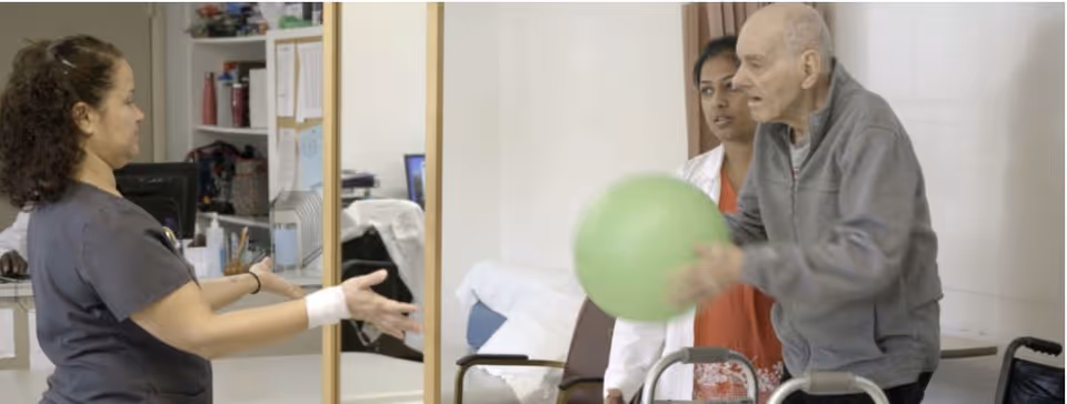 An elderly man holding a green exercise ball is assisted by a healthcare worker and a nurse in a rehabilitation center room with medical equipment and furniture in the background.