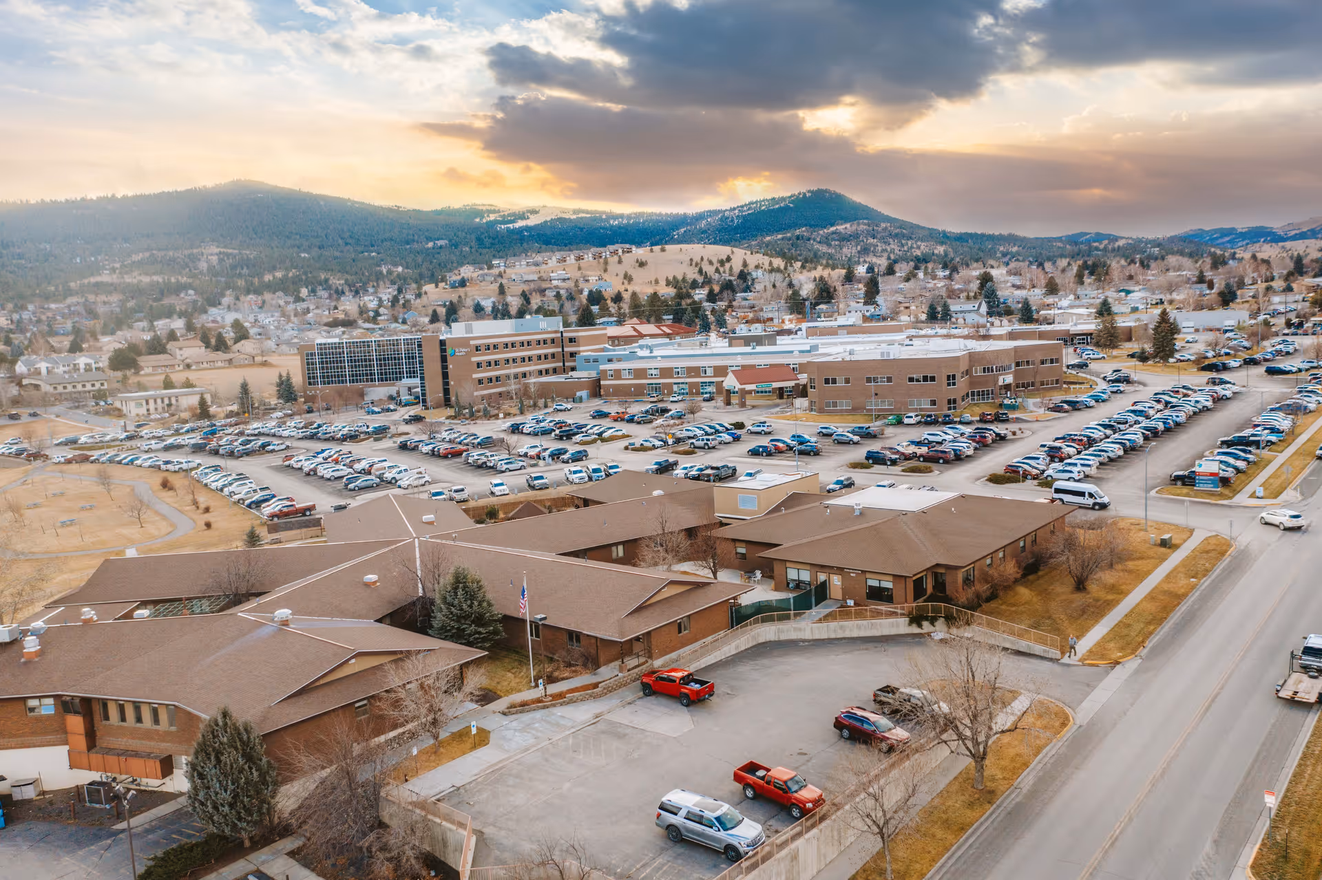 Aerial view of Cooney Healthcare and Rehabilitation facility with multiple connected buildings, large parking lots filled with cars, surrounding roads, and a backdrop of hills and a cloudy sky at sunset.