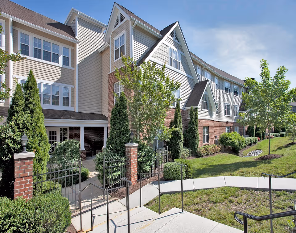 Front exterior of a multi-story senior living building with brick and siding, landscaped grounds, and a gated entrance.
