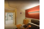 Interior view of a room in a healthcare facility with a bed, bedside table, and a red accent wall. The room has a door leading to a hallway with visible signage and lighting fixtures on the ceiling.