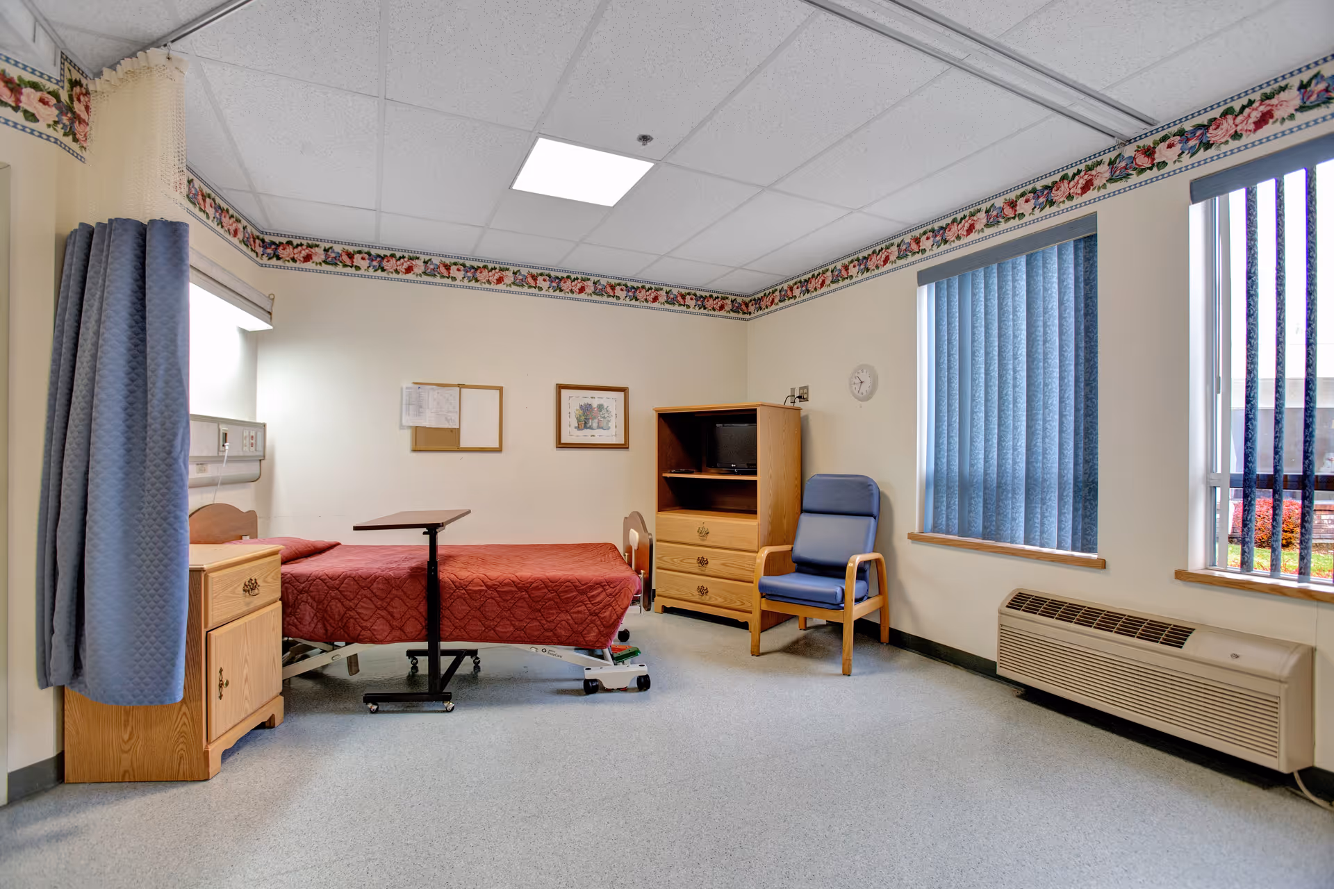 A simple, clean patient room in a senior living facility with a single bed covered in a red quilt, a wooden nightstand, an adjustable overbed table, a wooden cabinet with a small TV, a blue cushioned chair, and two windows with blue vertical blinds. The walls are light-colored with a floral border near the ceiling.