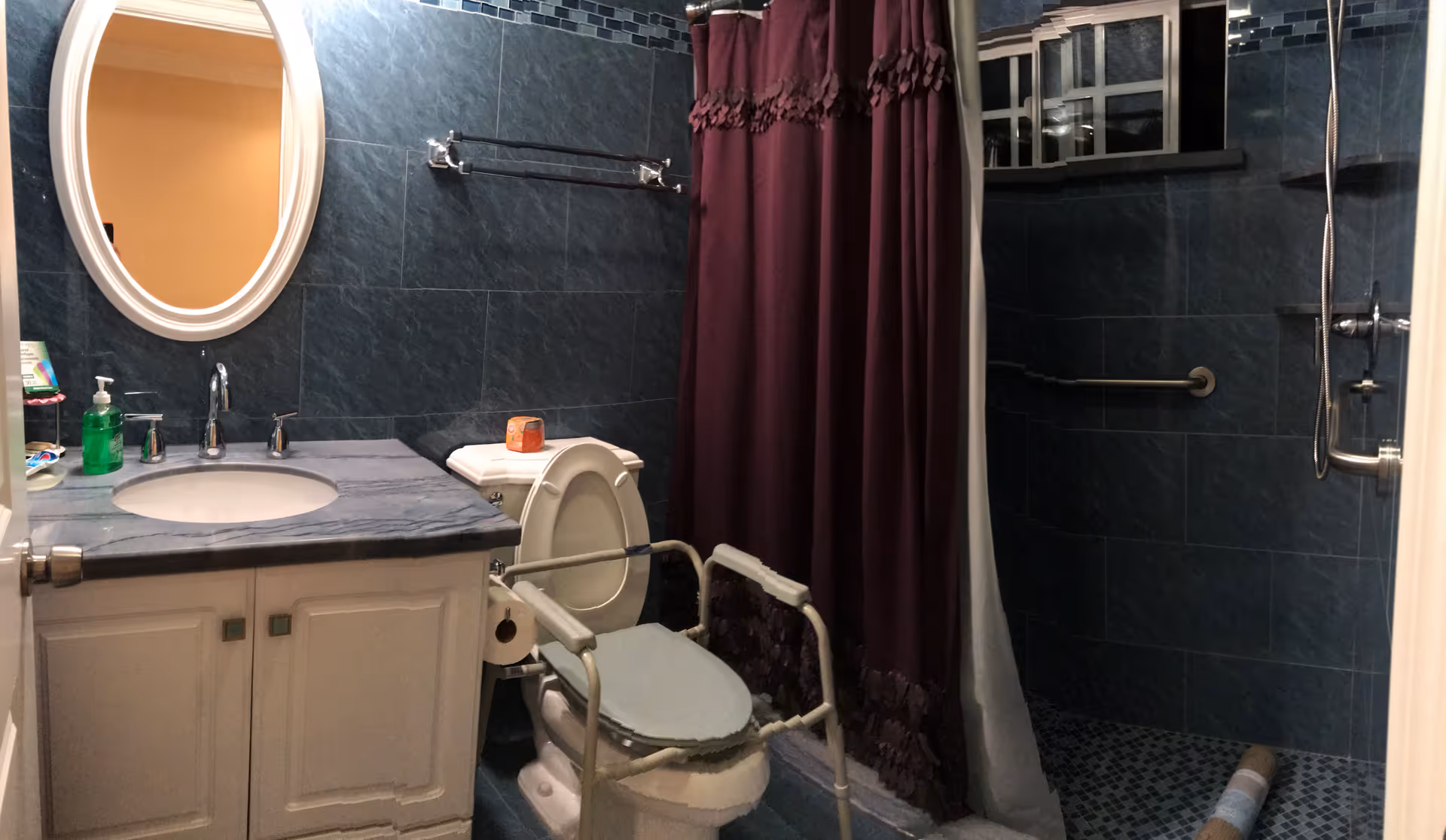 Bathroom with dark blue tiled walls and floor, a white vanity with a sink and oval mirror above it, a toilet with safety rails around it, a maroon shower curtain, and a walk-in shower with a grab bar and handheld showerhead.