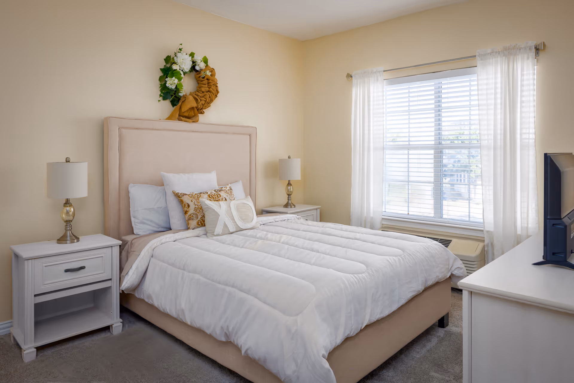 A neatly made bed with a beige upholstered headboard, white bedding, and decorative pillows including one with 'XO' on it. There are two white nightstands on either side of the bed, each with a lamp. A window with white curtains allows natural light into the room. A TV is placed on a white dresser opposite the bed. The walls are painted a soft beige color and a decorative wreath hangs above the headboard.