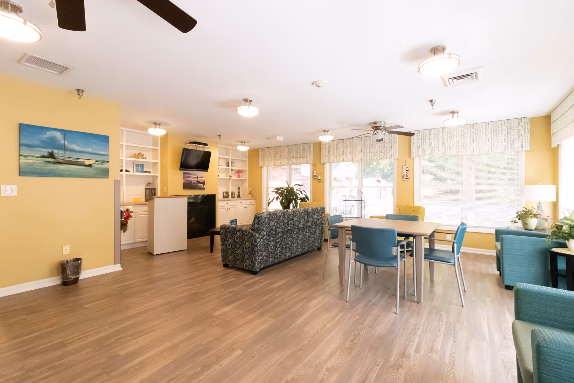 Bright communal lounge with sofas, chairs, a dining table, TV, and large windows.