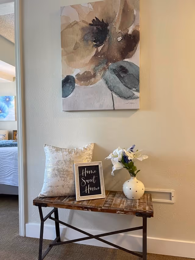 A small wooden bench with a decorative pillow, a framed sign that reads 'Home Sweet Home,' and a white vase with white and purple flowers. Above the bench hangs a large watercolor painting of abstract flowers in neutral tones. A bedroom with a bed and blue floral artwork is partially visible through an open doorway to the left.