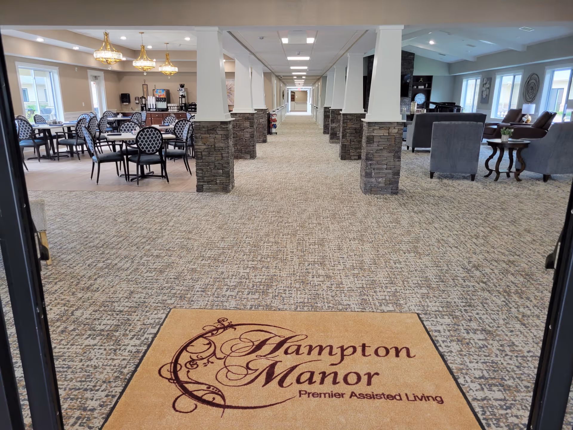 View from an entrance into a spacious interior of an assisted living facility. The foreground shows a beige floor mat with the text 'Hampton Manor Premier Assisted Living'. The area features a long carpeted hallway with stone pillars and white columns. To the left, there is a dining area with tables and chairs under elegant chandeliers. To the right, there is a lounge area with armchairs, side tables, and large windows allowing natural light.