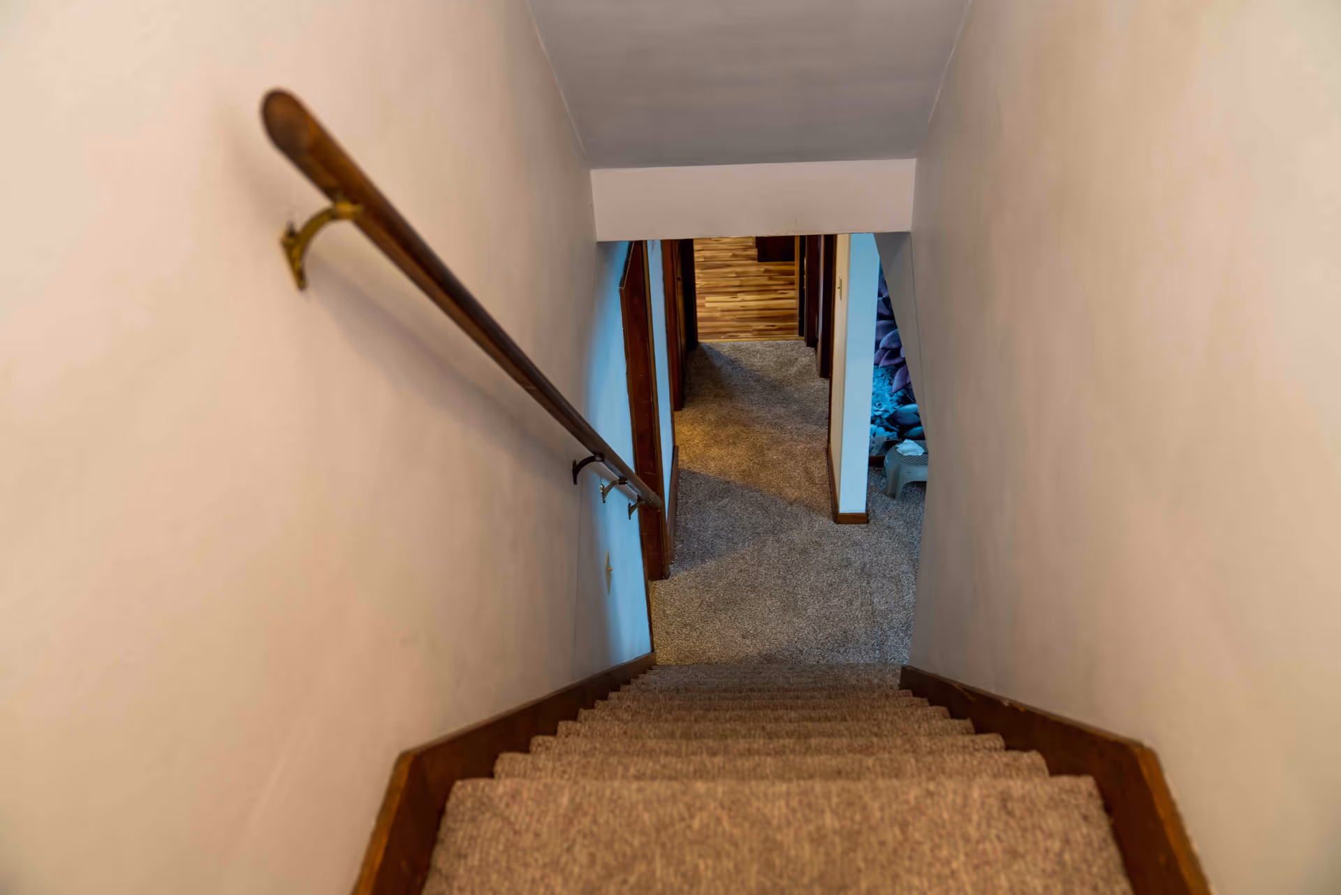 View looking down a narrow carpeted staircase with a wooden handrail leading to a carpeted hallway and open doorways.