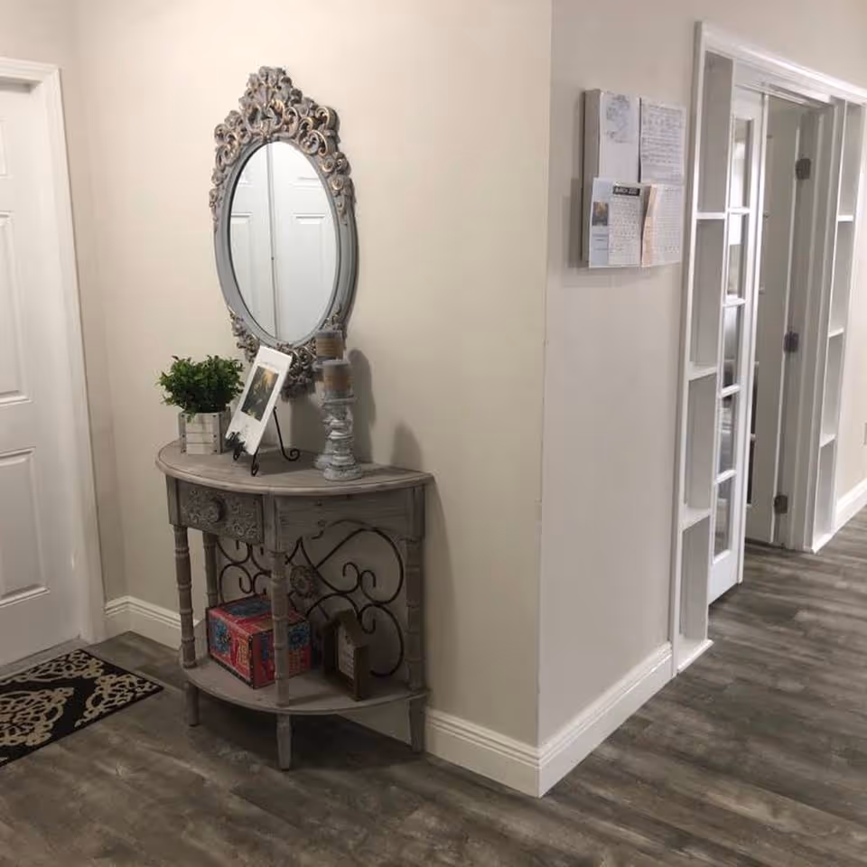 A hallway corner with a decorative wooden console table holding a small potted plant, a framed photo, and two candles on a stand. Above the table is an ornate oval mirror. The floor is wood-style laminate, and there are white doors and a bulletin board with papers on the wall.
