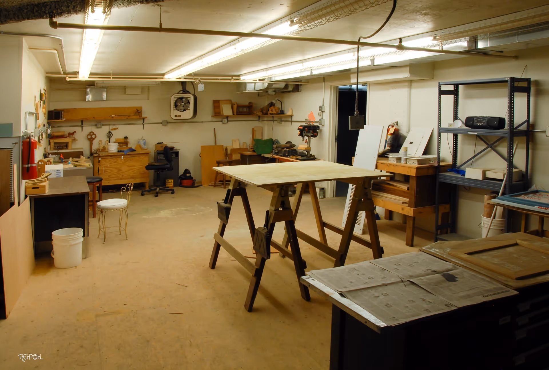 A spacious workshop room with wooden workbenches, tools, and equipment. The room has fluorescent ceiling lights, shelves with various items, a fan mounted on the wall, and a chair. The floor is wooden and there are various woodworking materials and supplies scattered around.