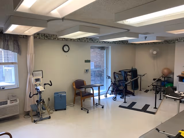 A therapy or rehabilitation room with exercise equipment including a stationary bike, parallel bars for walking practice, a walker, and a chair with a small table. The room has white walls, a clock, a window with curtains, and fluorescent ceiling lights.