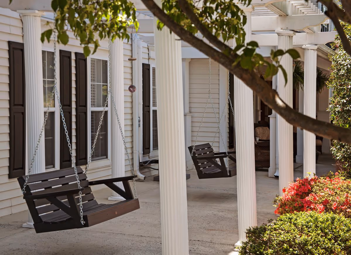 Outdoor porch area with two dark wooden swing benches hanging from a white pergola supported by white columns. The porch is adjacent to a building with white siding and dark window shutters. There are green bushes and red flowers in the foreground, and tree branches partially frame the top of the image.
