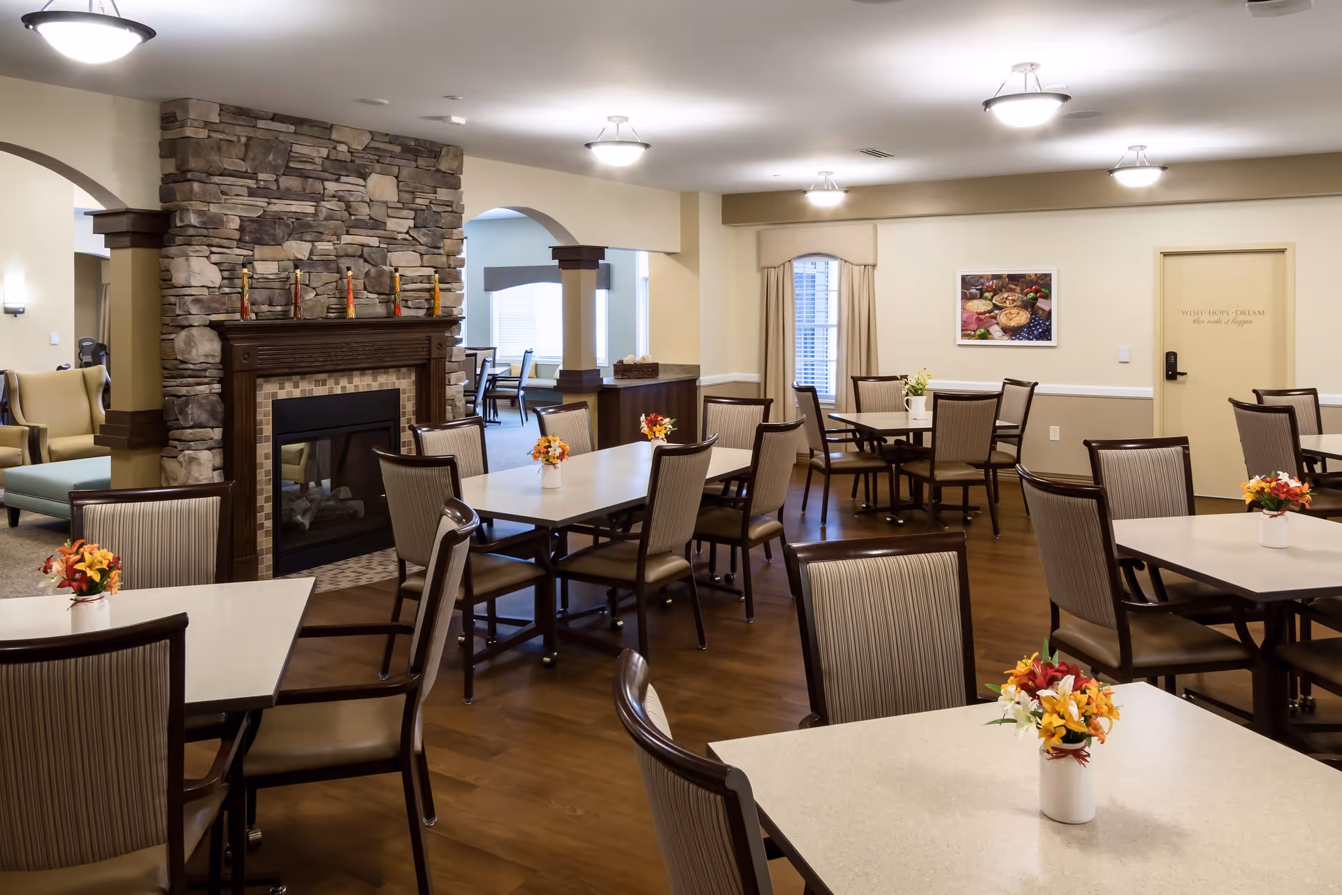 A spacious dining room with multiple tables and chairs arranged neatly. Each table has a small vase with colorful flowers. There is a stone fireplace with decorative items on the mantel. The room has warm lighting, wooden floors, and cream-colored walls with a framed picture and a door in the background.