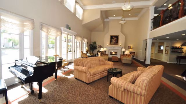 Bright, spacious senior living common room with striped sofas, a grand piano, large windows, and a fireplace.