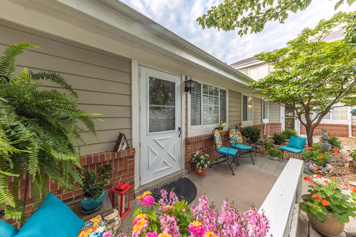 Outdoor patio area at Fort Scott Presbyterian Village featuring a white door with a wreath, two chairs with colorful cushions, various potted plants and flowers, a small table, and a tree providing shade.