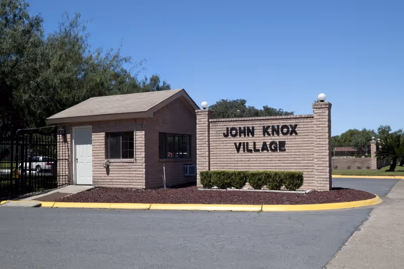 Entrance gatehouse and sign for John Knox Village, a brick structure with black lettering, surrounded by landscaping and a clear blue sky.