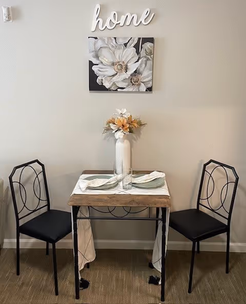 A small dining area with a square wooden table set for two, featuring two black metal chairs with decorative backs. On the table is a white vase with flowers and two place settings with napkins and glasses. Above the table on the wall is a decorative sign that says 'home' and a painting of white flowers.