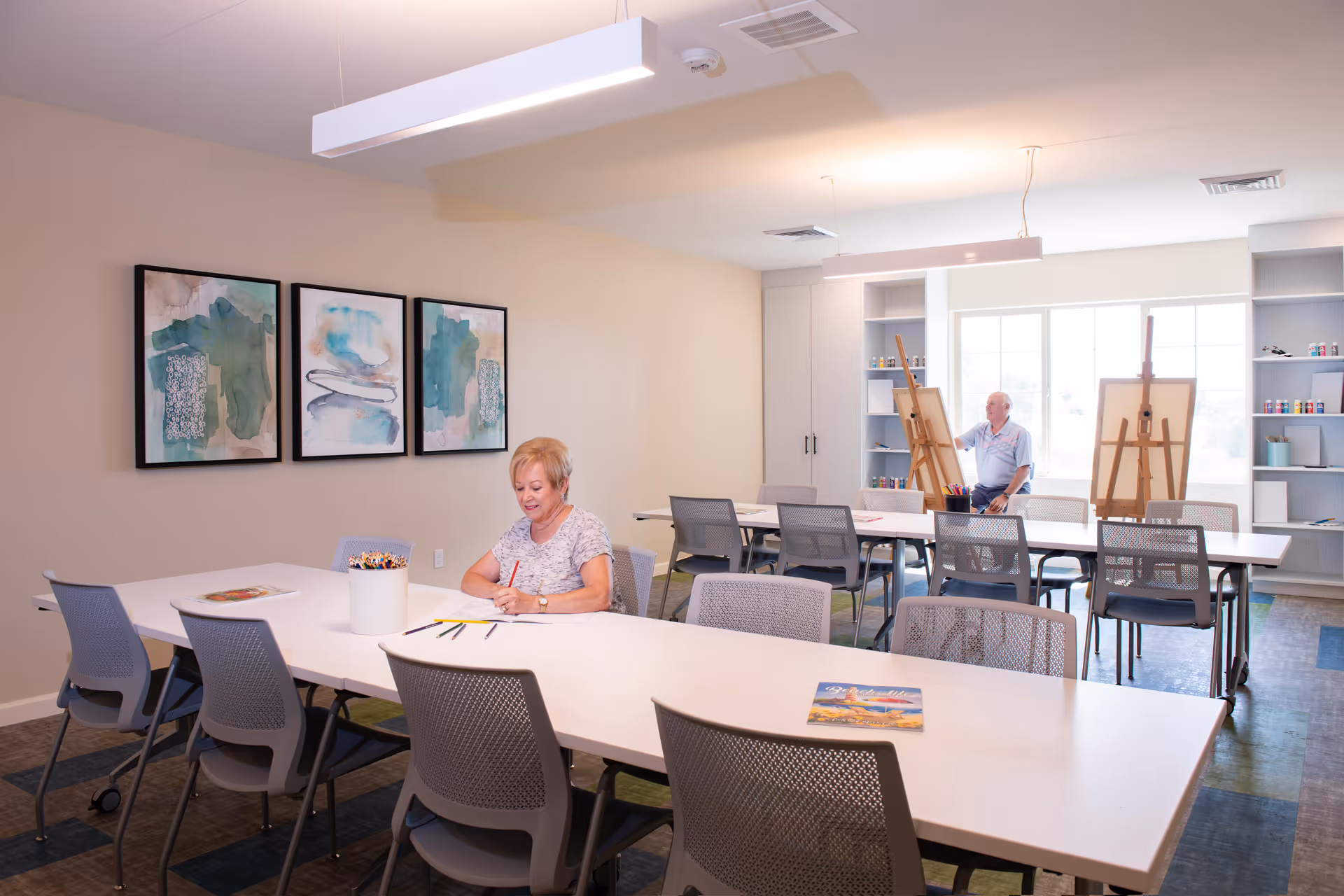 A bright art room with two elderly individuals engaged in creative activities. One woman is sitting at a white table drawing with colored pencils, while a man is seated near two easels by a large window, possibly painting. The room has modern chairs, abstract artwork on the wall, and shelves with art supplies.