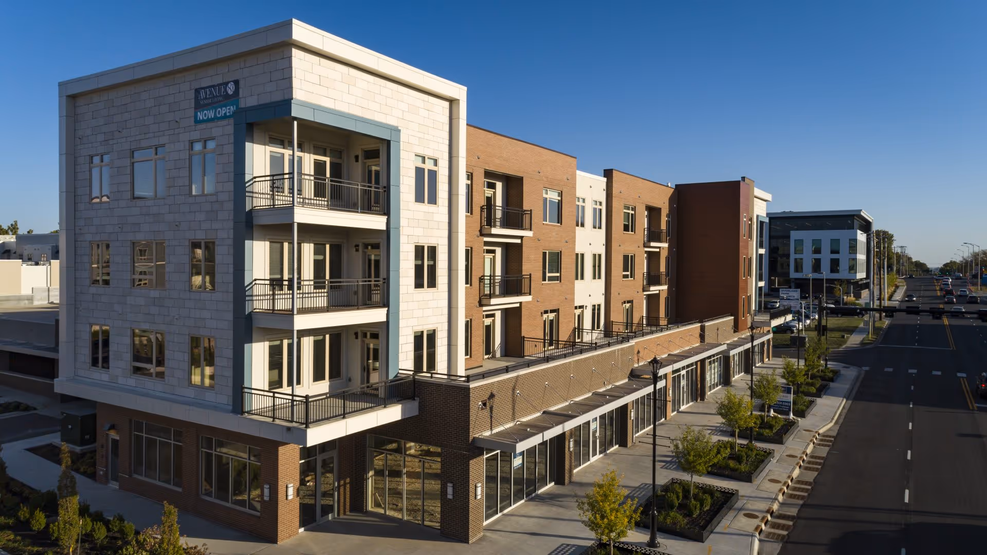 Exterior view of a modern multi-story building with balconies and large windows, located along a wide street with trees and street lamps. The building has a sign that reads 'Avenue 81 Senior Living NOW OPEN'.