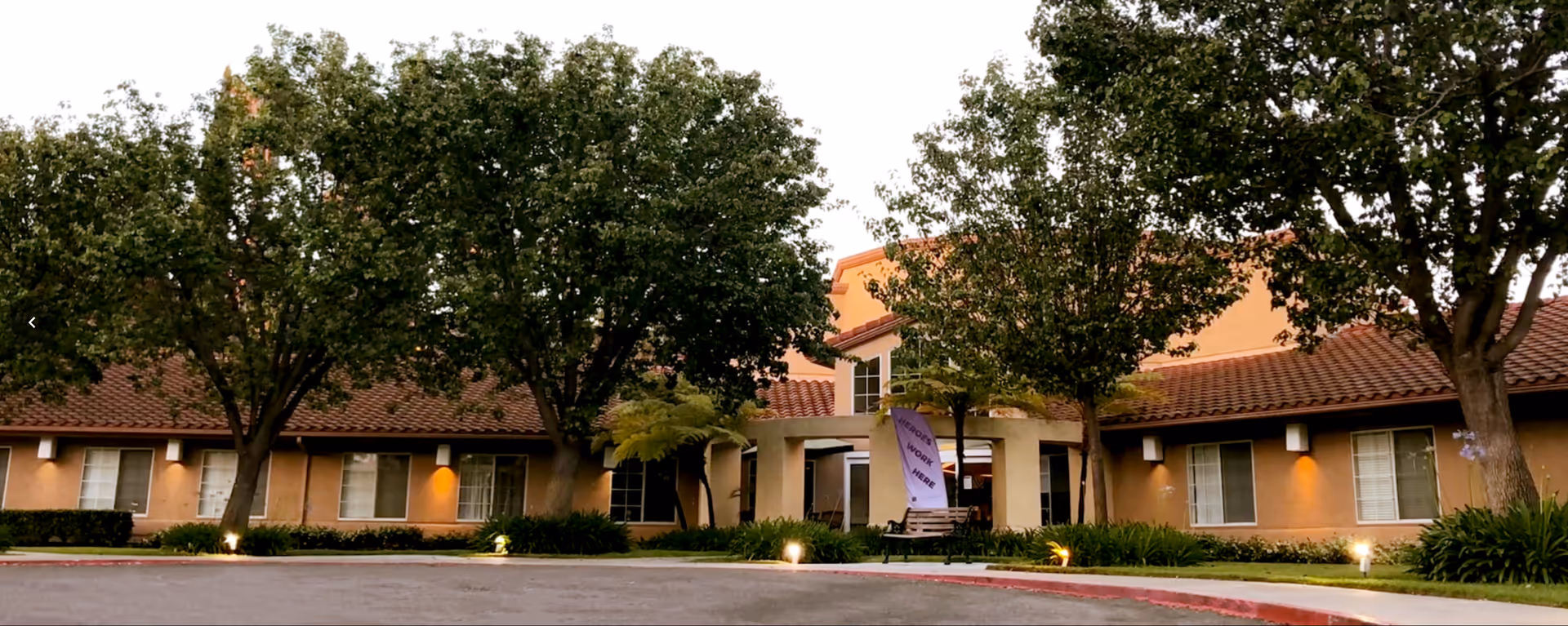 Exterior view of Villa Valencia Healthcare Center showing a single-story building with a tiled roof, surrounded by trees and landscaped greenery, with a paved driveway in front.