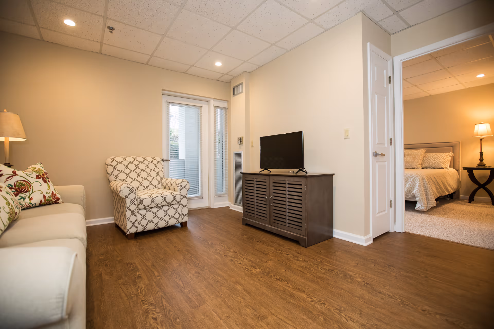 A cozy living room area with a beige sofa adorned with floral cushions, a patterned armchair, a wooden TV stand with a flat-screen television, and a doorway leading to a bedroom with a bed, nightstand, and lamp. The room has wooden flooring and beige walls with recessed lighting in the ceiling.