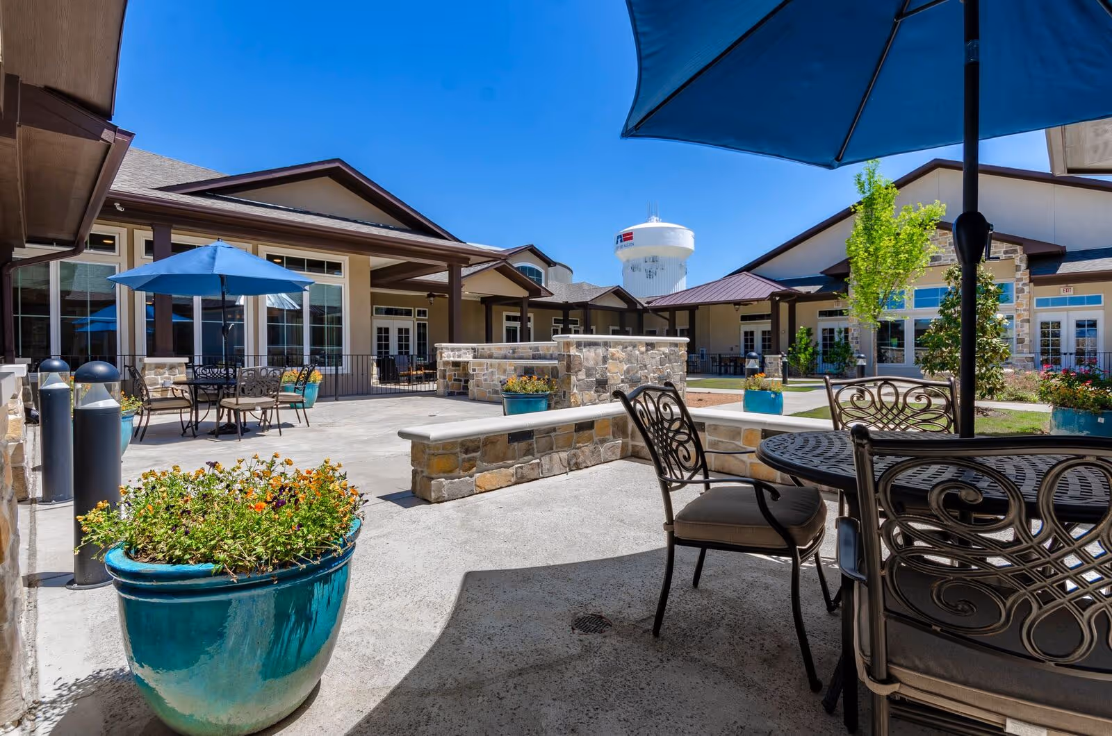 Outdoor courtyard area at Grand Brook Memory Care of Allen at Twin Creeks featuring patio tables with umbrellas, cushioned chairs, stone walls, potted plants with flowers, and a clear blue sky with a water tower visible in the background.