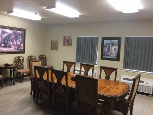 Long wooden dining table surrounded by chairs in a well-lit room with framed artwork and windows with blinds.