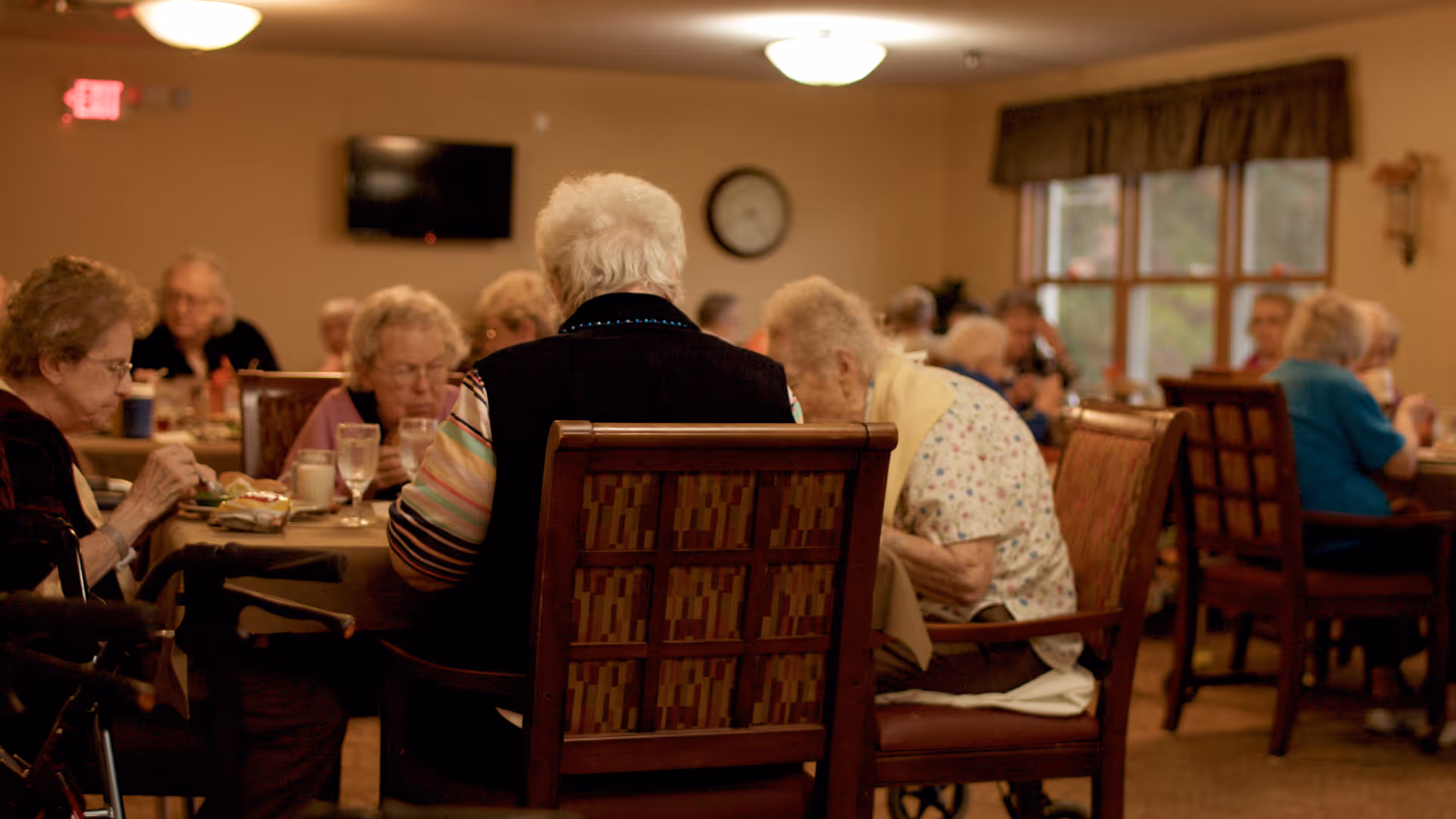 A group of elderly people sitting at tables in a dining room, eating and socializing. The room has warm lighting, a wall clock, a television mounted on the wall, and large windows with curtains.