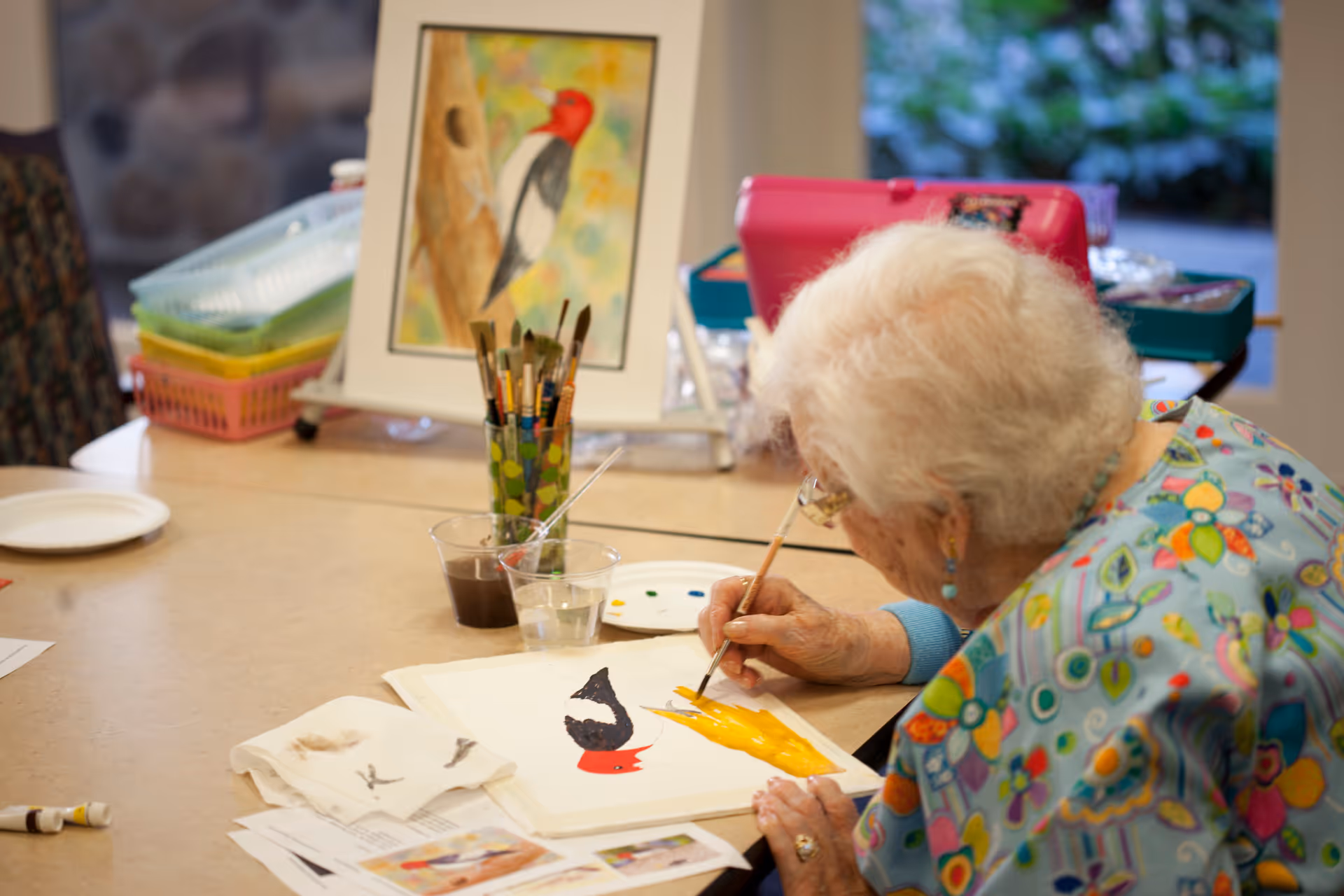 An elderly woman with white hair and colorful clothing is seated at a table, painting a bird on a piece of paper. Various painting supplies, including brushes in a cup, paint cups, and a reference picture of a woodpecker, are on the table. The setting appears to be a bright indoor room with a window showing greenery outside.
