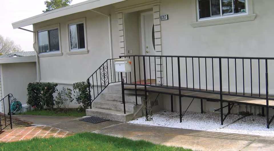 Exterior view of a single-story building with a white door, windows, and a black metal ramp with handrails leading to the entrance. There are concrete steps beside the ramp, a white mailbox mounted on the ramp railing, some bushes near the building, and a green garden hose coiled on the side. The ground has a mix of grass, concrete, and white gravel.