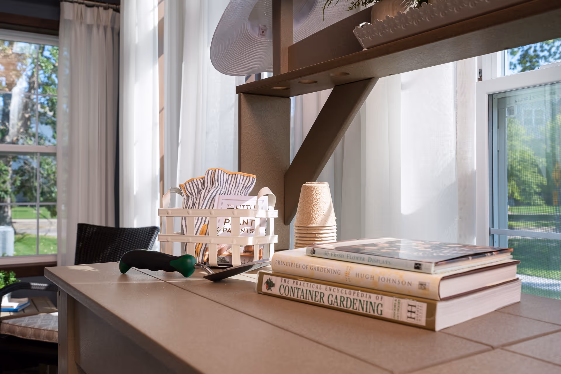 A sunlit indoor gardening station with gardening books, a small white basket containing gardening gloves, a small green-handled gardening tool, and a stack of disposable plant pots. The background shows large windows with white curtains and a view of green trees outside.
